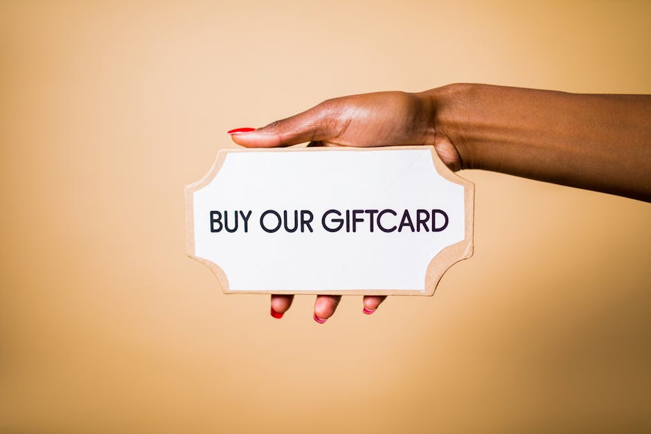 referral program incentives - Close-up of a hand holding a 'Buy Our Giftcard' sign on a brown backdrop.