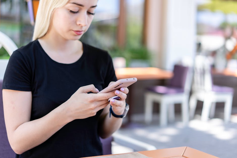 SMS referral marketing - Young woman in a black shirt texting on her smartphone at an outdoor cafe.