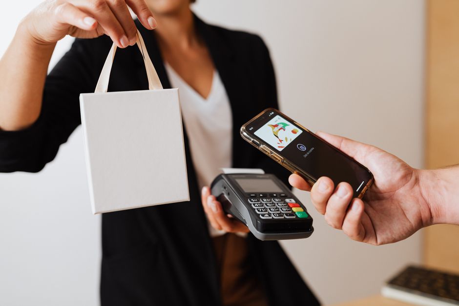 affiliate marketing news - A person uses a smartphone for contactless payment with a payment terminal, while receiving a shopping bag.